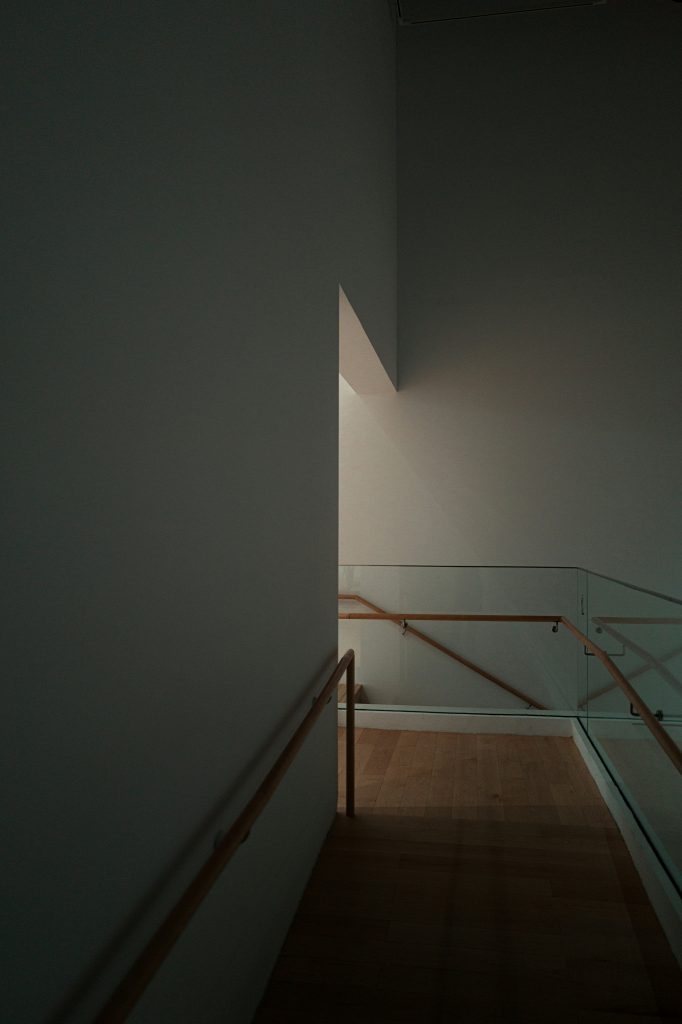 Image of a dim corridor, with white walls and a glass half wall on the right leading to a left bend through an arch on the left wall. There is light tastefully pouring through the left arch. Shot by De an Sun.